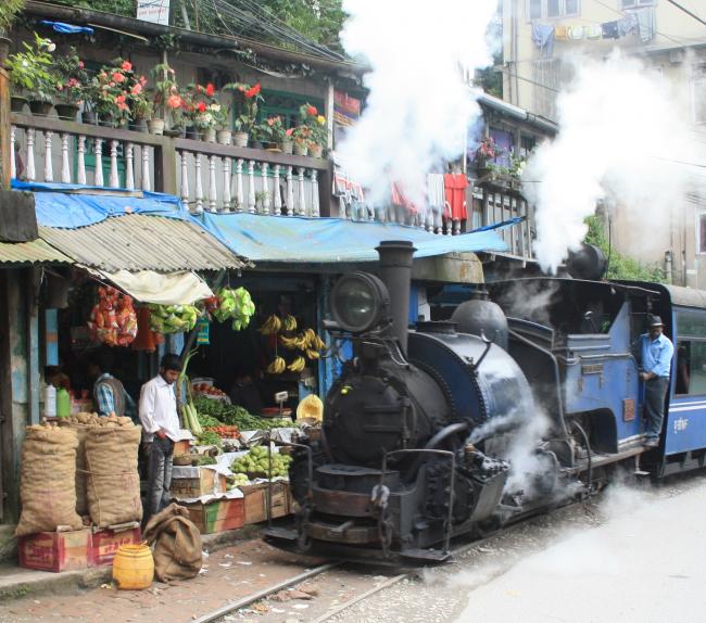 Photo description and credit: A steam train to Darjeeling about 5 minutes before arriving at its final destination, passing by a fruit and vegetable shop. Arne Hückelheim – Wikipedia Commons
